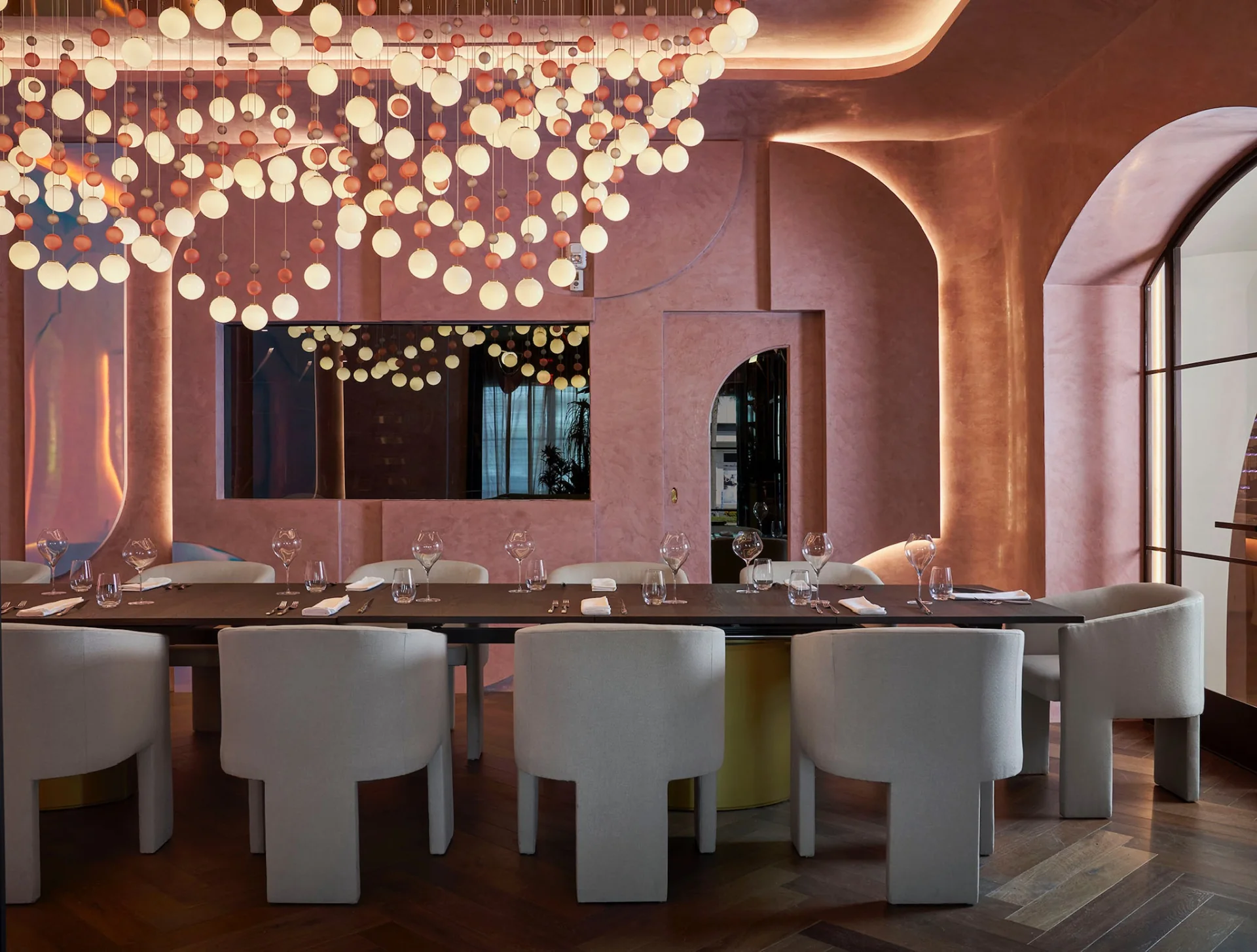 Composed dining room at Cocody — sculptural pendant lighting drops over a long table set with fine glassware, all held within warm terracotta plaster walls.