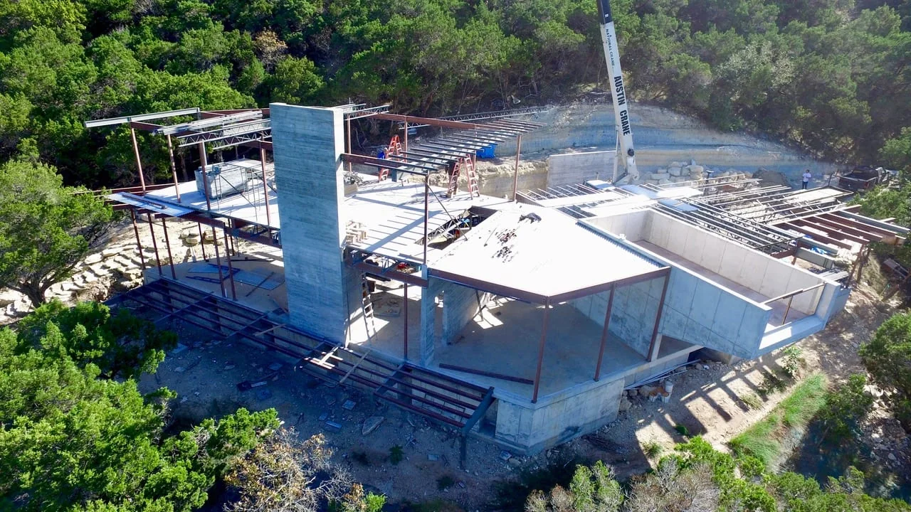 A modern home under construction — steel frame, concrete walls, and crane on a hillside site.